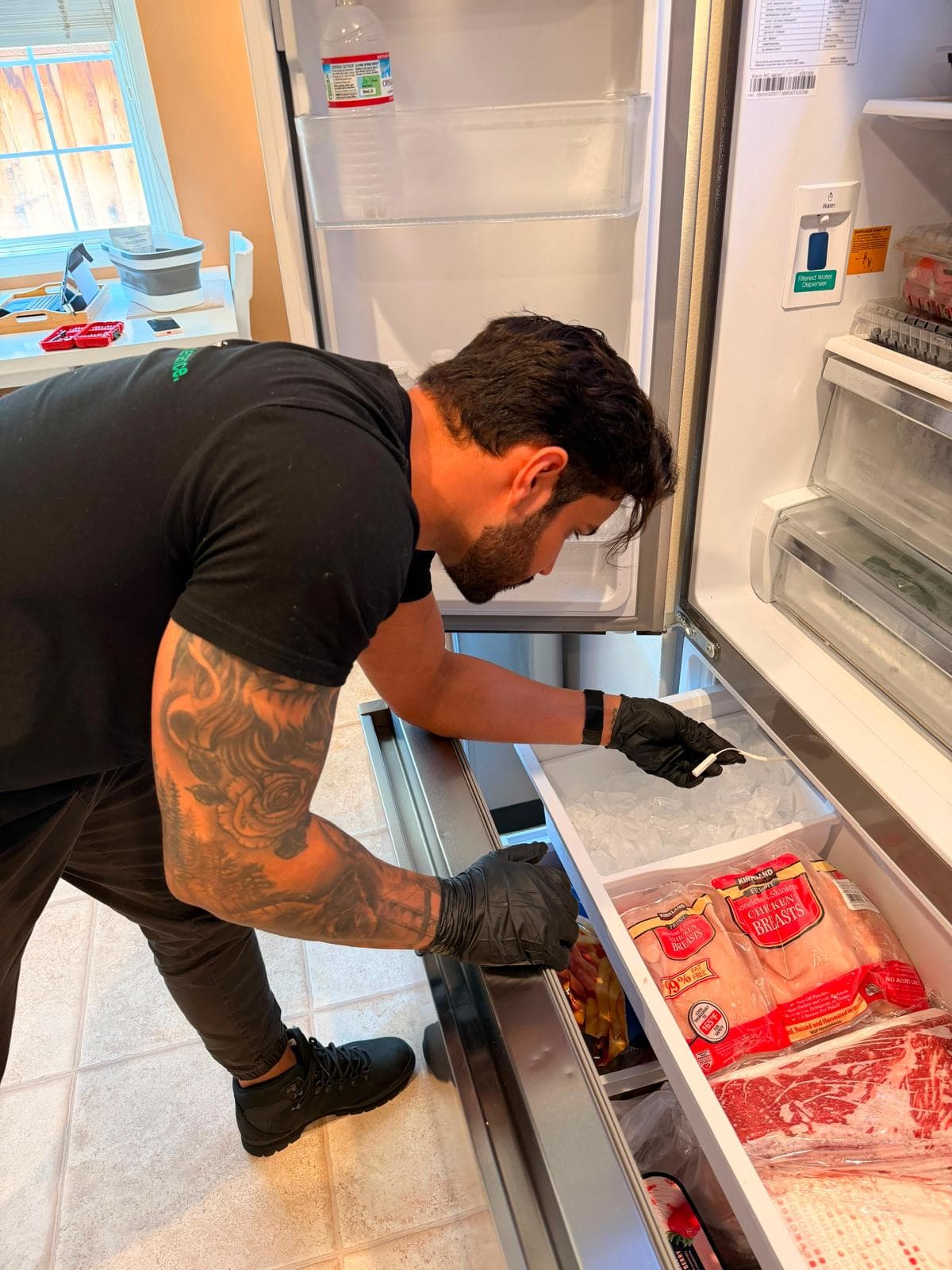 A man with arm tattoos and black gloves inspects the freezer drawer of a refrigerator.