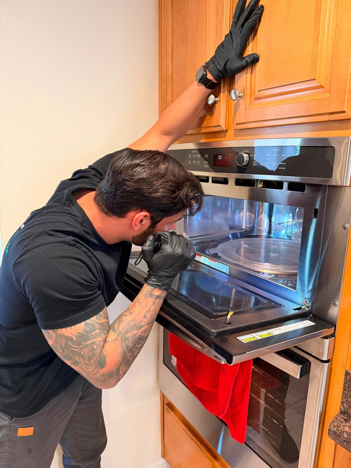 Tattooed technician wearing black gloves inspects the interior of a built-in kitchen microwave oven.