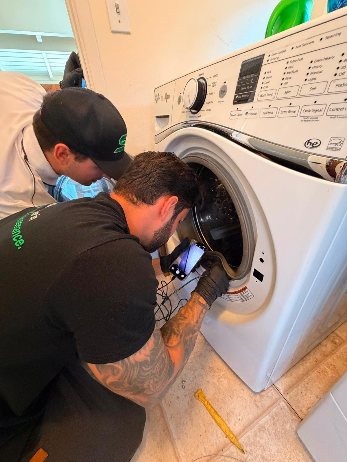Two technicians inspect the interior of a white front-loading washing machine using a smartphone camera.